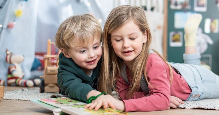 Zwei Kinder, ein Junge und ein Mädchen, liegen nebeneinander auf dem Boden und blättern gemeinsam in einem Buch. Im Hintergrund ist Spielzeug zu sehen.