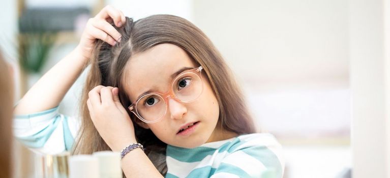 Mädchen mit Brille untersucht sorgenvoll ihren Haaransatz vor dem Spiegel.