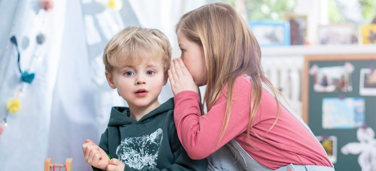 Ein blondes kleines Kind in einem dunkelgrünen Pullover schaut erstaunt, während ein Mädchen mit langen braunen Haaren ihm ins Ohr flüstert. Beide Kinder sitzen in einem hellen Raum.