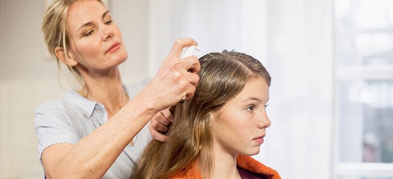 Eine Frau sprüht NYDA® Läusemittel in das Haar eines Mädchens mit langen braunen Haaren, das ein orangefarbenes Handtuch über den Schultern trägt. Das Mädchen sitzt ruhig, während die Behandlung auf die Kopfhaut aufgetragen wird.