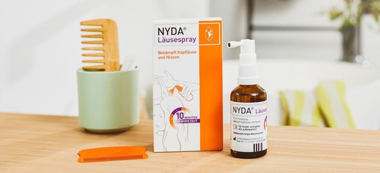 NYDA® Läusespray mit Verpackung, Flasche und orangefarbenem Läusekamm auf einem Holztisch, im Hintergrund ein Holzkamm in einem Becher.