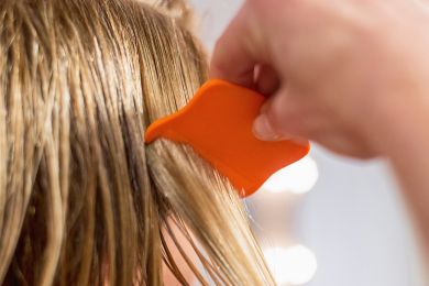 Nahaufnahme einer Hand, die einen orangefarbenen Läusekamm durch das nasse, blonde Haar eines Kindes zieht. Der Läusekamm hat eine breite, flache Form und wird vorsichtig durch eine Haarpartie geführt, um Läuse und Nissen zu entfernen. Im Hintergrund ist ein unscharfer, heller Bereich zu erkennen, der den Fokus auf den Kamm und die Haare lenkt.