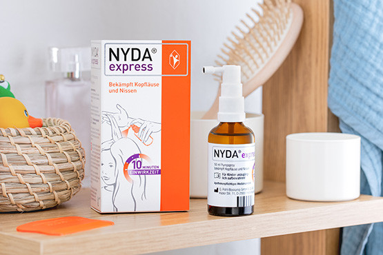 NYDA® express Flasche und Verpackung zur Bekämpfung von Kopfläusen und Nissen auf einem Regal im Badezimmer, neben einem orangefarbenen Läusekamm und weiteren Pflegeutensilien.