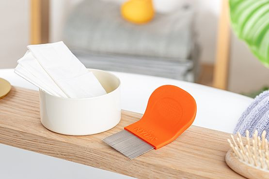 NYDA® Läusekamm – effektives und schonendes Auskämmen von Läusen und Nissen. Orangefarbener NYDA® Läuse- und Nissenkamm liegt neben einem Becher auf einer Holzablage, umgeben von weiteren Pflegeutensilien.