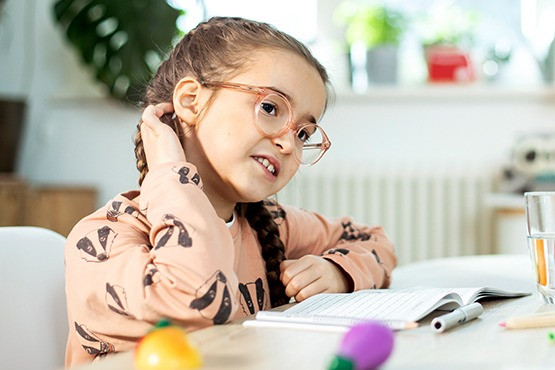 Ein Mädchen mit Brille und geflochtenen Zöpfen sitzt an einem Tisch und kratzt sich am Hinterkopf. Vor ihr liegen ein Notizbuch und bunte Spielzeuge. Im Hintergrund sind Pflanzen und ein helles Fenster zu sehen.