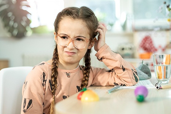 Ein Mädchen mit Brille und langen, geflochtenen Zöpfen sitzt an einem Tisch und kratzt sich mit einem genervten Gesichtsausdruck am Kopf. Vor ihr liegen bunte Spielzeuge und ein Glas Wasser.