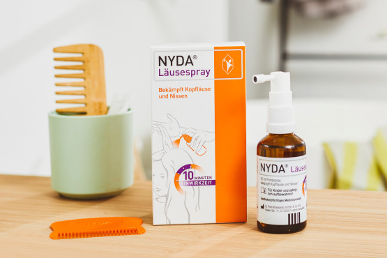 NYDA® Läusespray mit Verpackung, Flasche und orangefarbenem Läusekamm auf einem Holztisch, im Hintergrund ein Holzkamm in einem Becher.